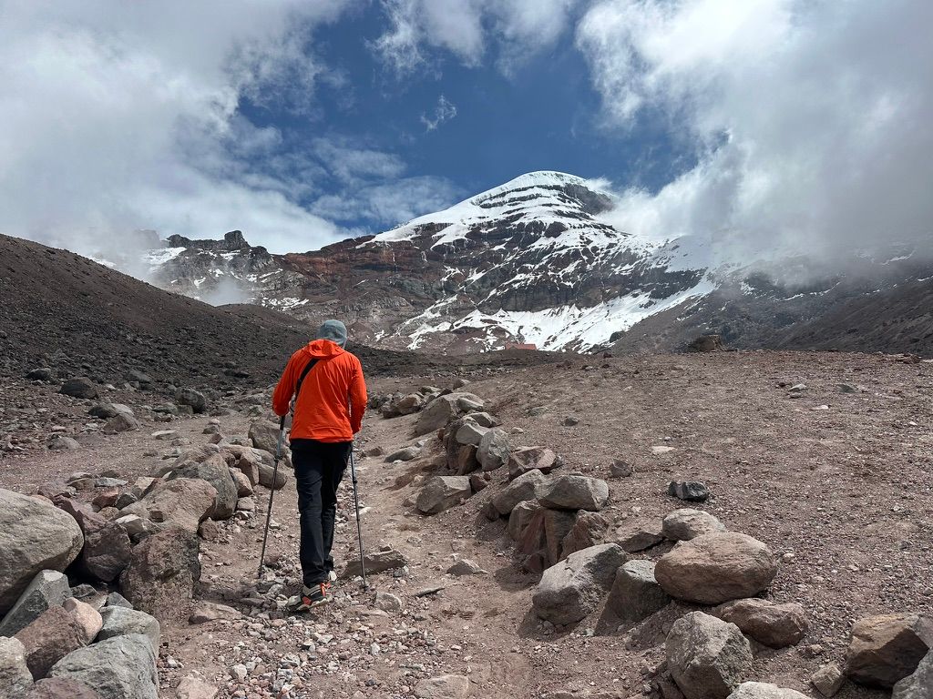 A trekker with poles on a rocky Andes trail approaching the snow-capped summit of Chimborazo under dramatic clouds